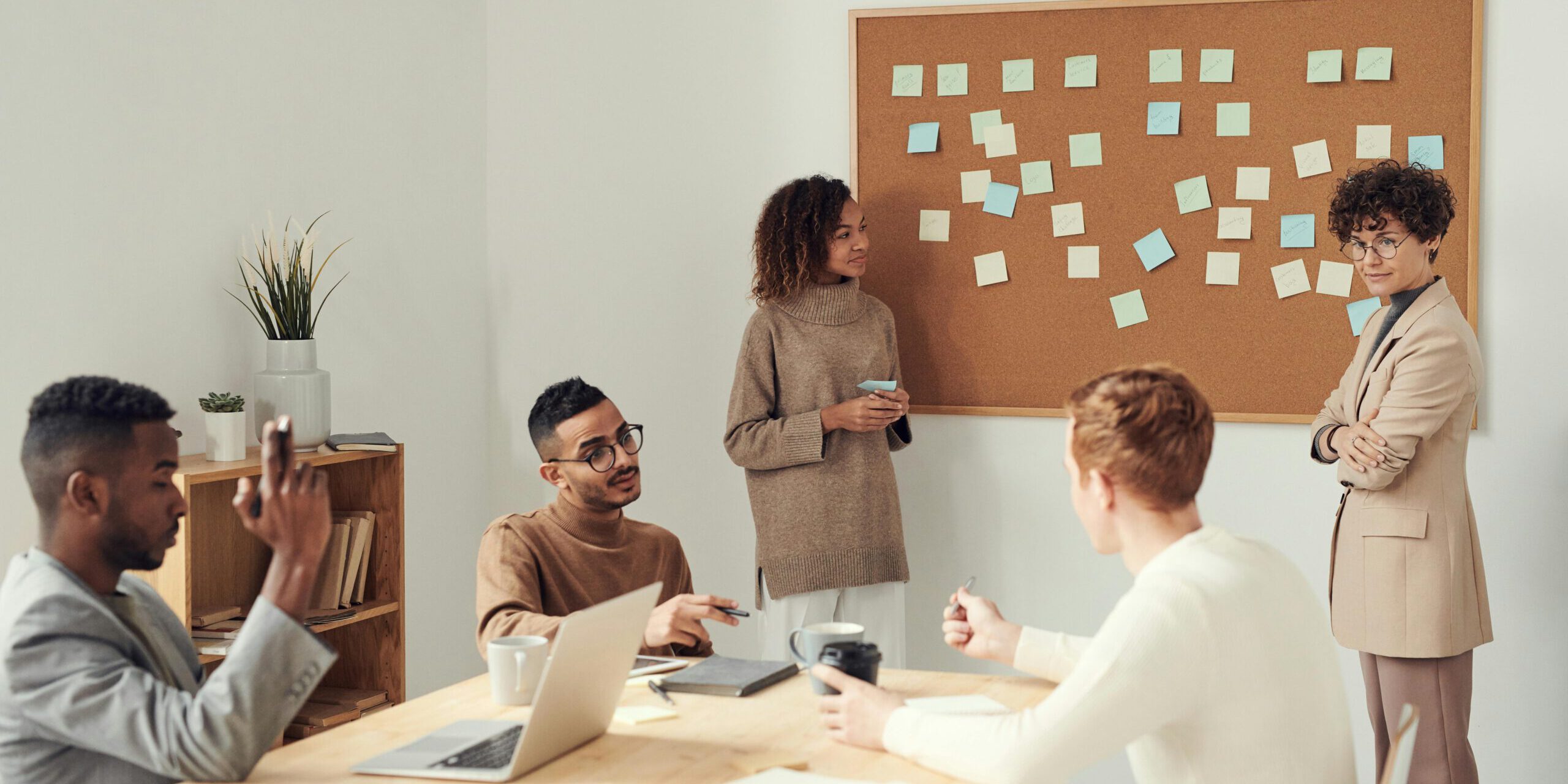 Taakherschikking vijf professionals zitten en staan rondom een tafel terwijl ze in overleg zijn. Er staat een laptop op tafel en twee professionals zijn bezig met een groot bord met post-its